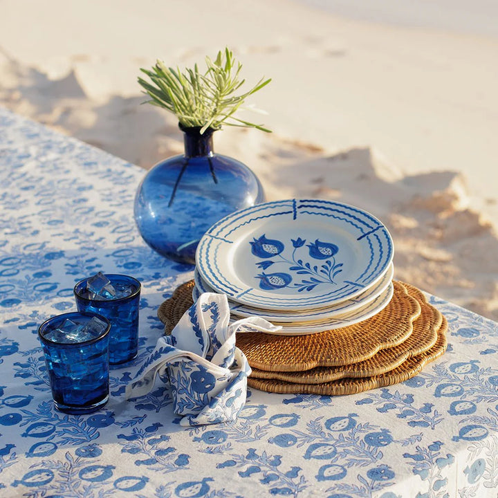 Table setting with blue and white floral plates, glasses, and woven coasters on a patterned tablecloth.