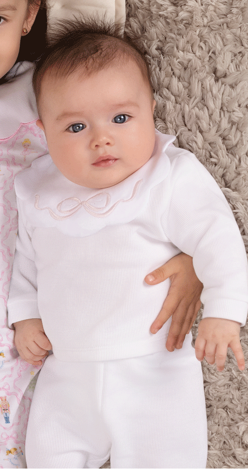 A baby and a toddler lying side by side on a textured beige rug. The toddler wears a light pink long-sleeve pajama with a white collar and a small fairy pattern, while the baby wears a white long-sleeve top and matching white pants.