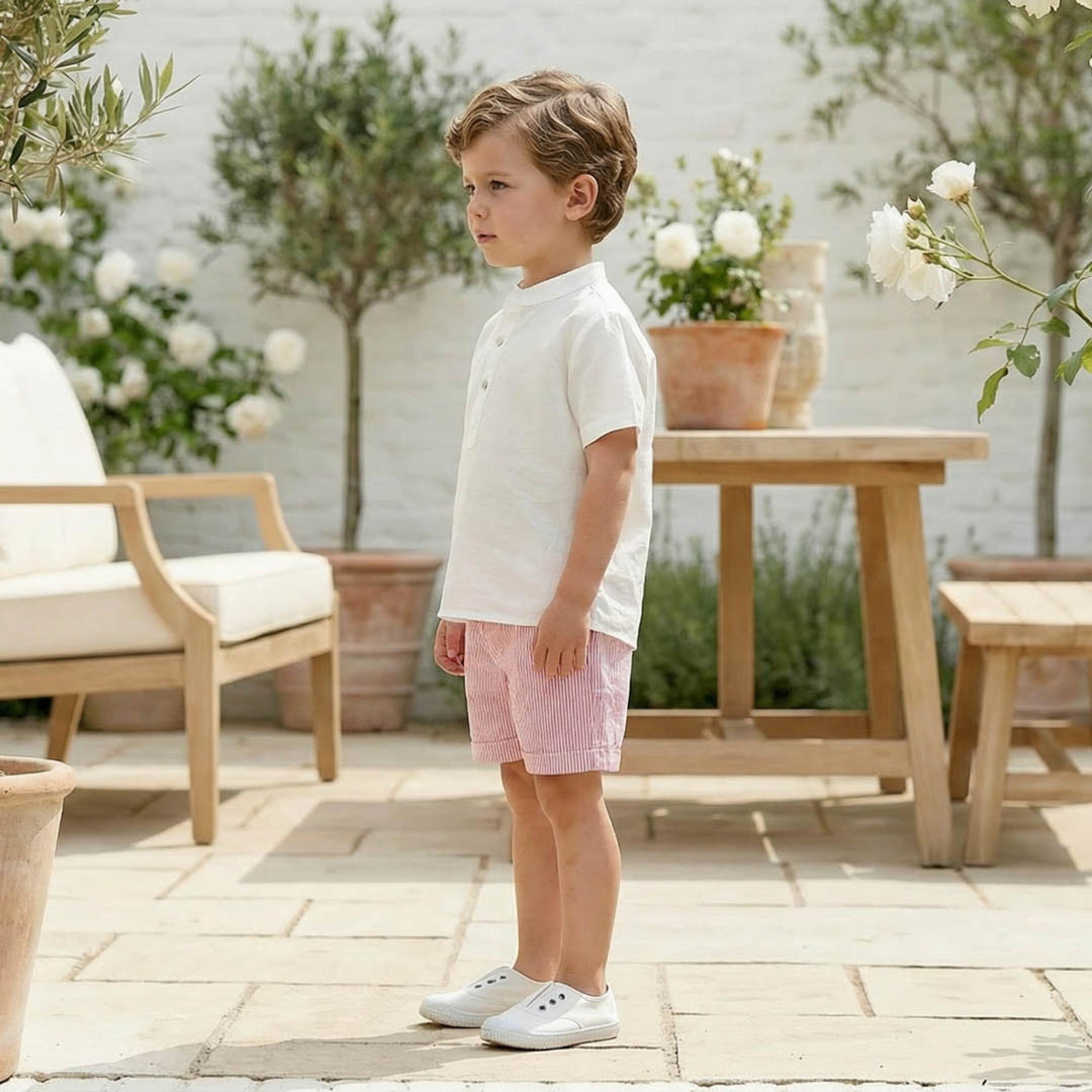 Child in a white shirt and red shorts standing on a patio with outdoor furniture and plants.