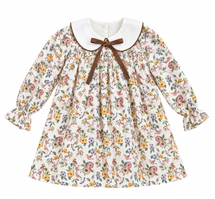 A long-sleeve girls dress with a dense multicolor floral print on an off-white background, featuring a contrasting white Peter Pan collar with a brown trim and a matching brown bow at the neckline.
