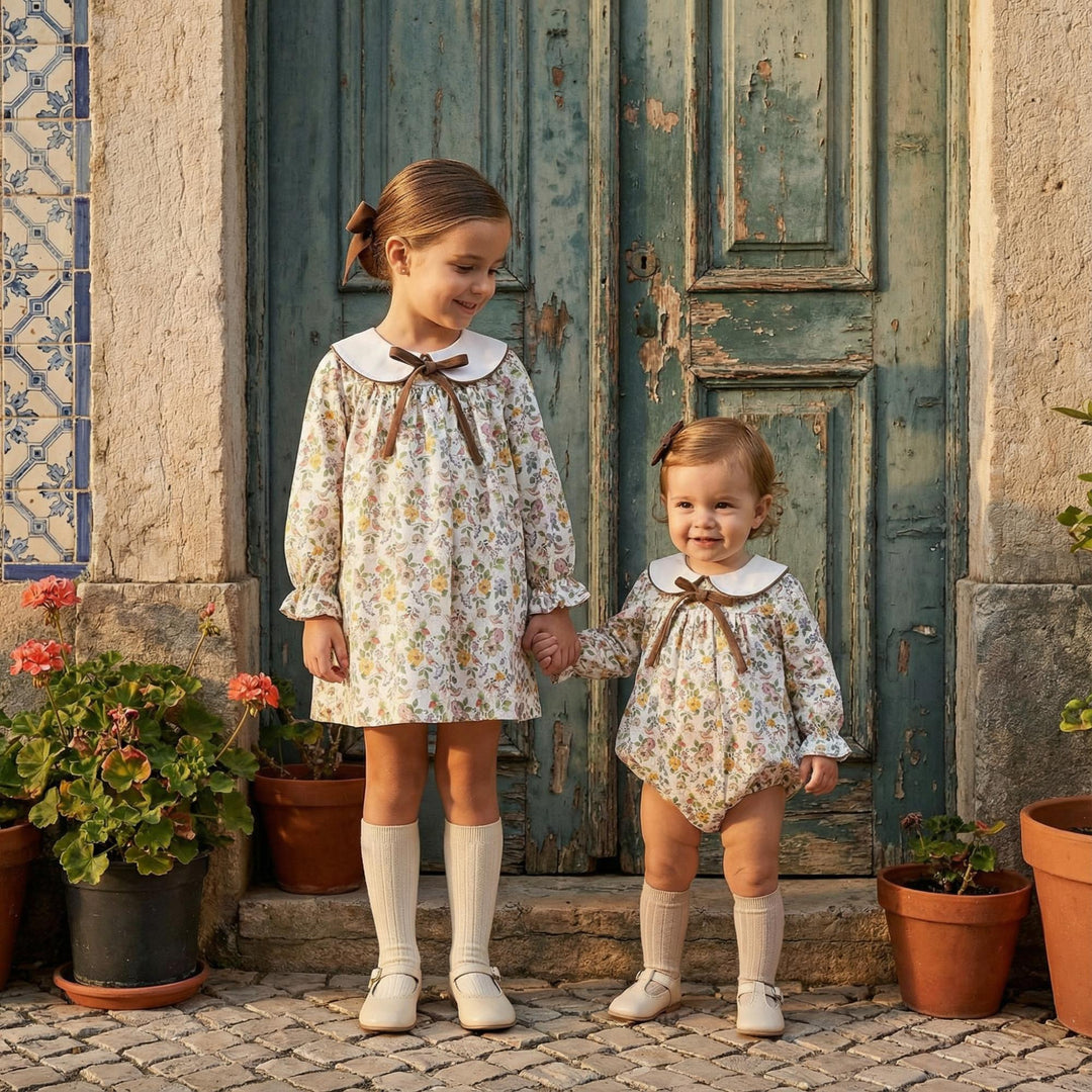 Two young girls standing hand in hand in front of a weathered teal wooden door, wearing matching floral long-sleeve dresses with white Peter Pan collars and brown bows. The older girl wears a knee-length dress and white knee-high socks, while the younger girl wears a matching floral romper and white socks.