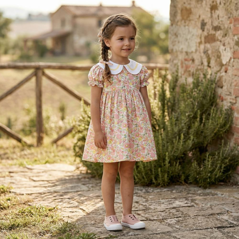 Young girl in a floral dress standing outdoors near a rustic wall and wooden fence.