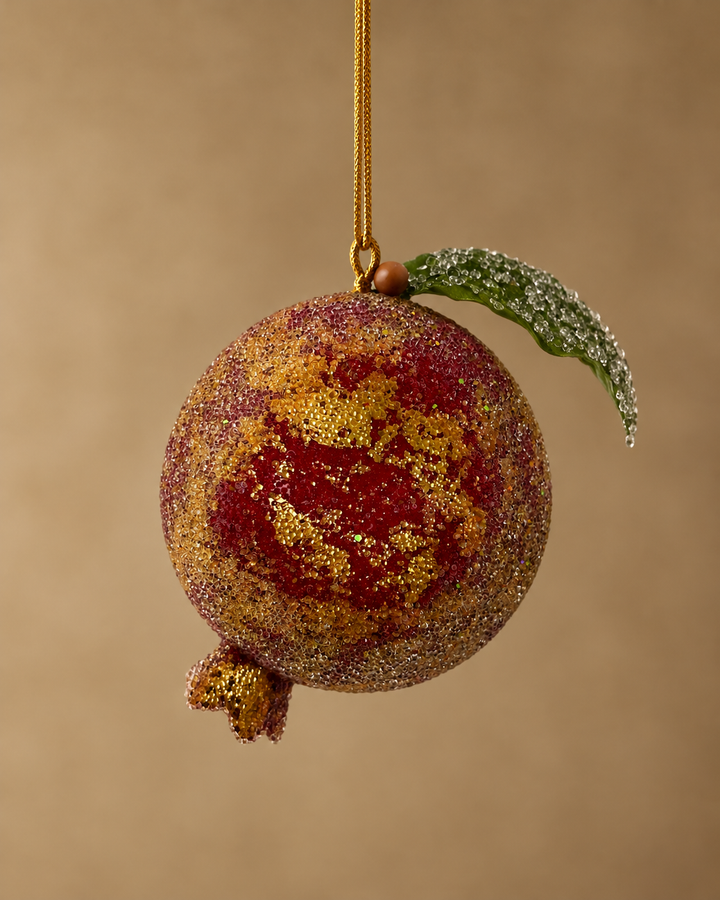 A decorative hanging ornament shaped like a pomegranate, covered in textured red and gold glitter, with a small green leaf on a gold string, set against a solid warm brown background.
