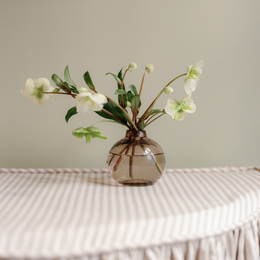A small round translucent brown glass vase holding white flowers with green leaves, placed on a cream striped fabric-covered surface against a plain light beige wall.