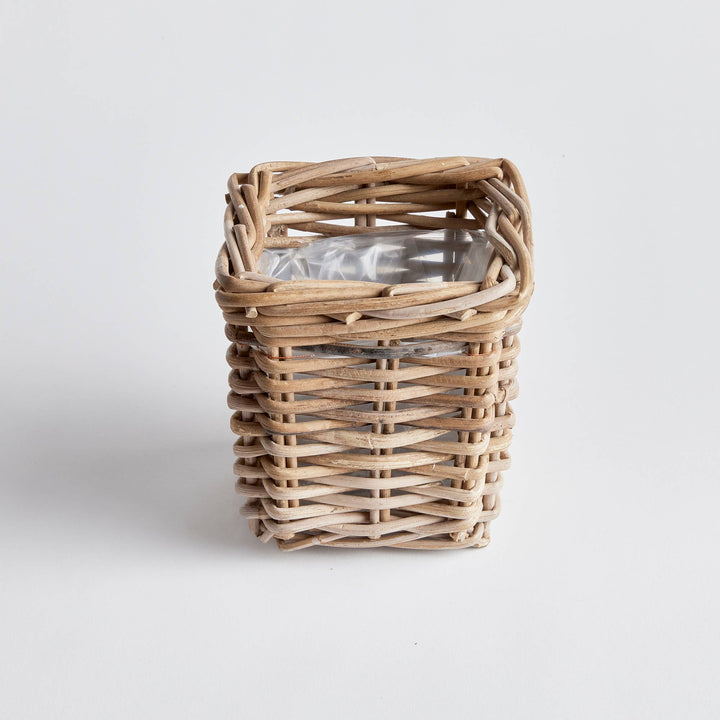 A small square woven wicker basket with a clear plastic wrap on a light gray background.