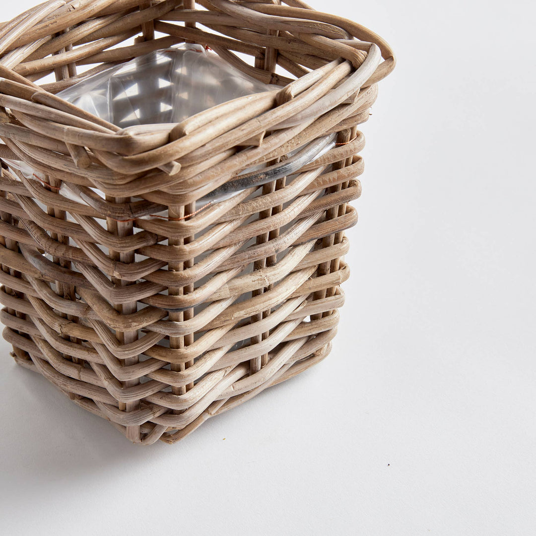 A close-up of a square woven wicker basket made from natural light brown reeds, containing a clear glass container, set against a plain white background.