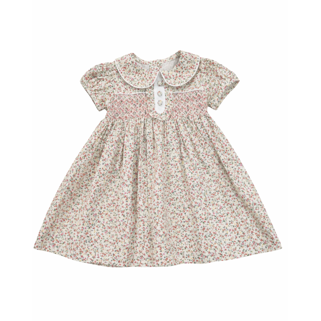 A baby girl's short-sleeve dress with a white Peter Pan collar, featuring a delicate multicolor floral print on a cream background and a smocked chest panel with a white button closure.