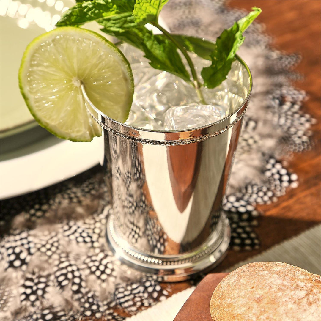 Silver mint julep cup with a lime wedge and mint leaves on a patterned tablecloth.