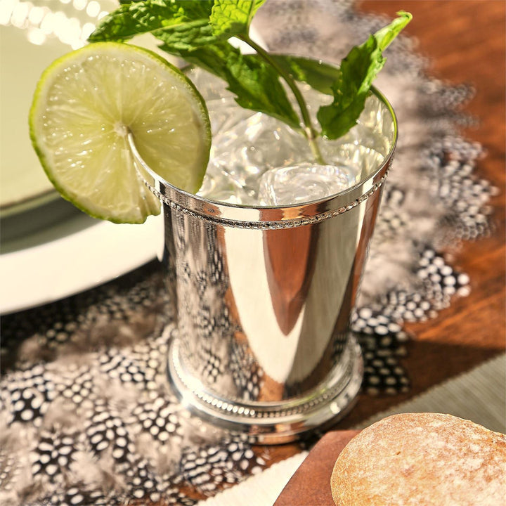 Silver mint julep cup with a lime wedge and mint leaves on a patterned tablecloth.