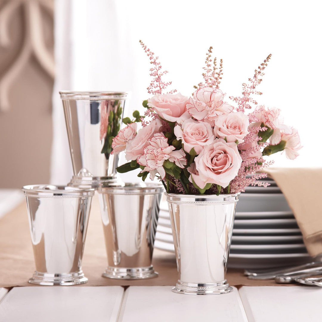 Three silver metal bucket-style cups arranged on a tabletop beside a pink floral centerpiece in a clear cup, with plates and a napkin in the softly blurred background.