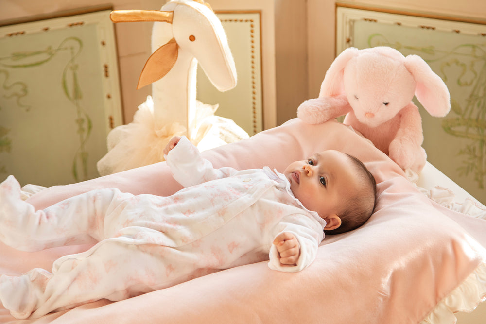 A baby in a white footed onesie lies on a pink changing pad in a nursery, with a pink plush bunny toy and a white swaddle blanket nearby.