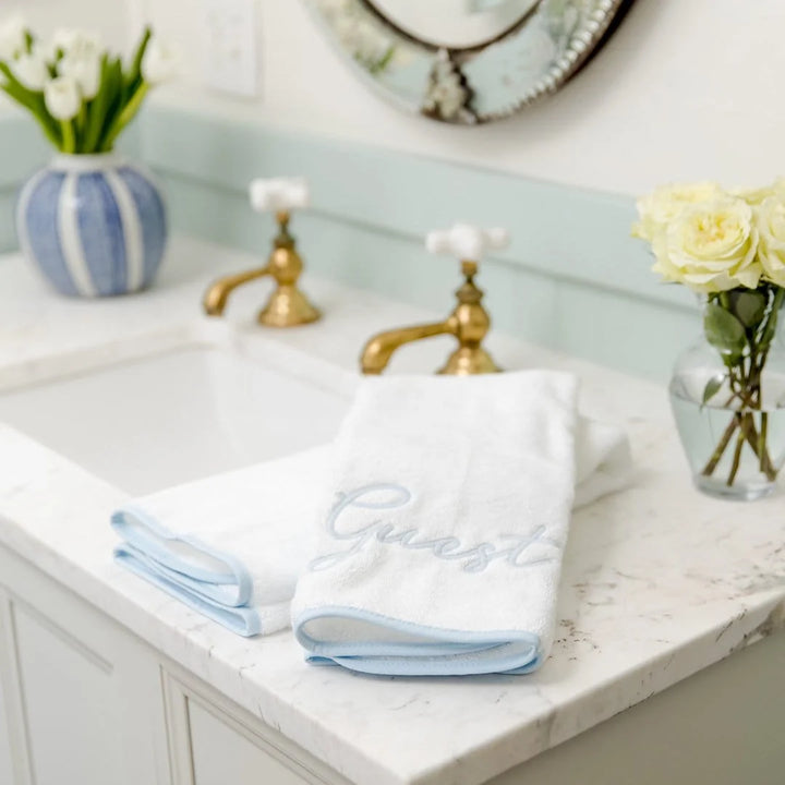 White towels with blue trim on a bathroom counter with flowers and a mirror.