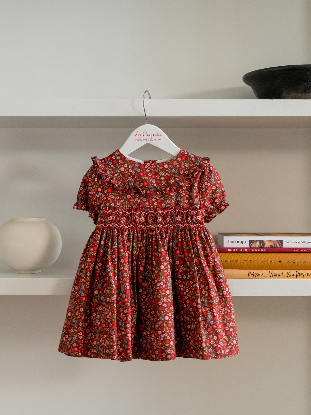 A toddler girl's short-sleeve dress with a red and green floral print, featuring a smocked waist and ruffled neckline, hanging on a white hanger from a bookshelf.