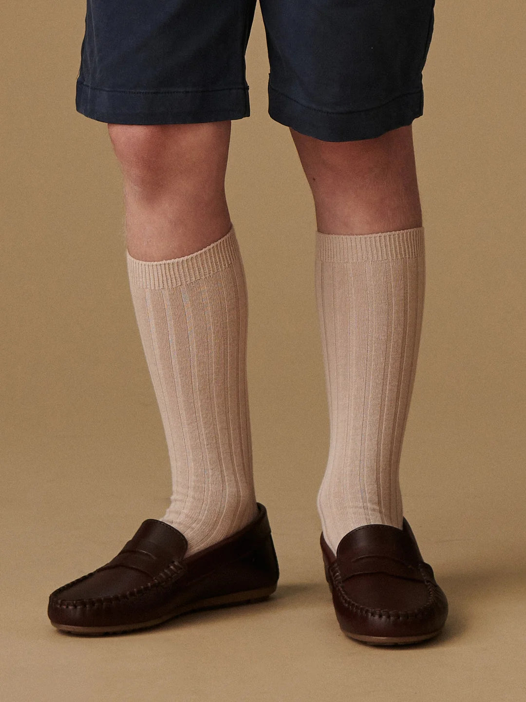 A person wearing beige ribbed knee-high socks with dark brown leather loafers and navy blue shorts, standing against a solid tan background.