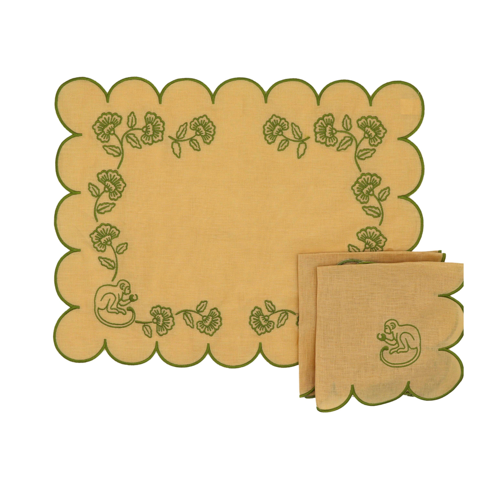 A set of natural linen table linens featuring scalloped edges and green embroidered details, including a large placemat and matching napkins with a monkey motif and floral border.