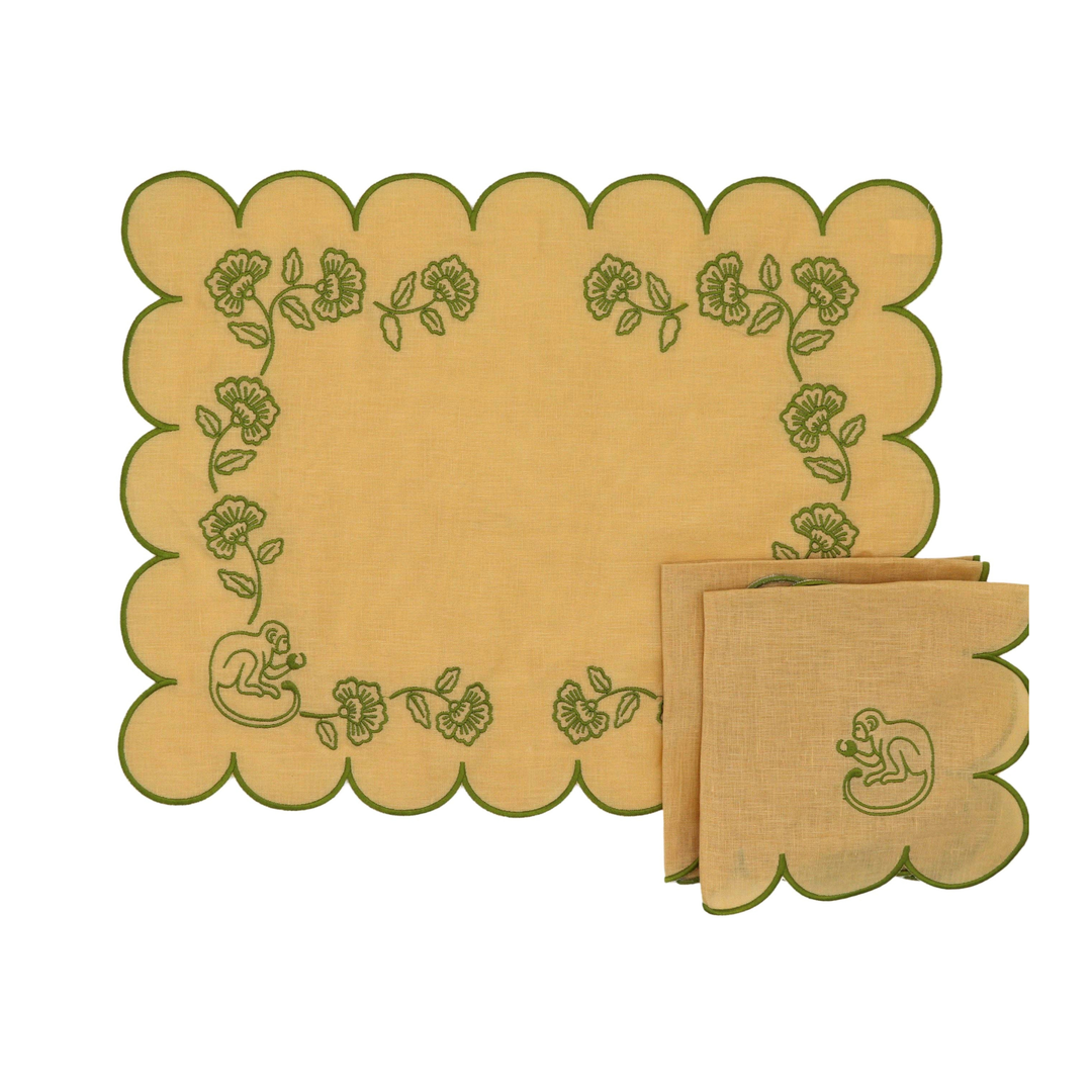 A set of natural linen table linens featuring scalloped edges and green embroidered details, including a large placemat and matching napkins with a monkey motif and floral border.