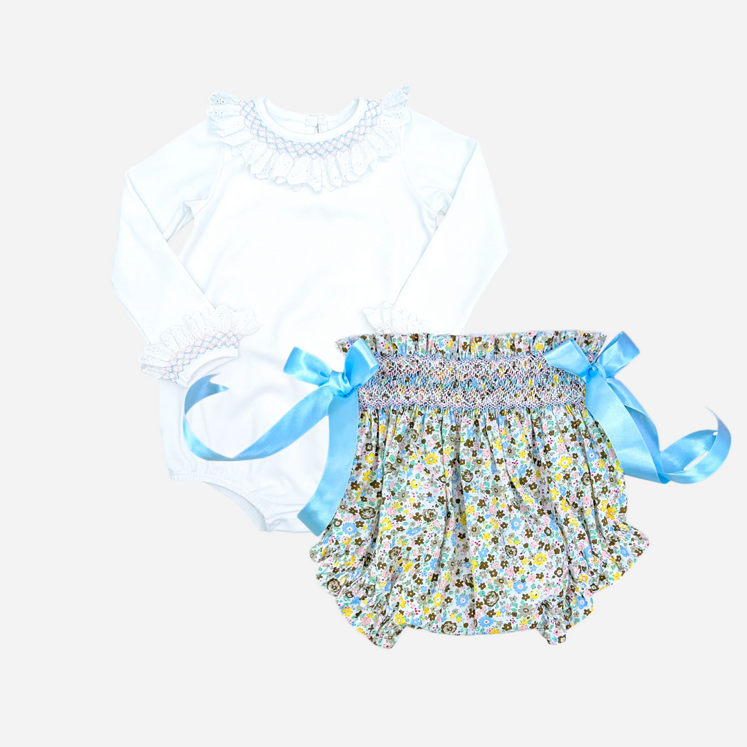 A two-piece baby outfit set featuring a white long-sleeve bodysuit with a ruffled mock neck collar, paired with smocked floral bloomers in a multicolor pattern with light blue satin bows.