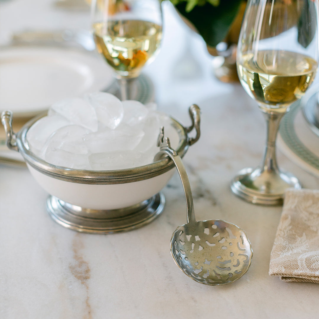 Silver ladle with decorative patterns on a marble surface, next to a bowl of ice and wine glasses.