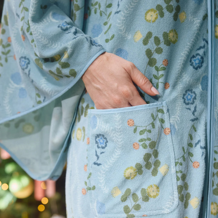 A close-up of a person wearing a light blue fleece robe with a floral vine pattern in green, yellow, and blue. The person's hand is tucked into a front patch pocket.