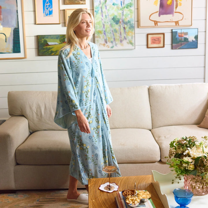 A woman wearing a light blue floral robe stands in a living room in front of a beige sofa, with framed artwork on the wall and a coffee table in the foreground.