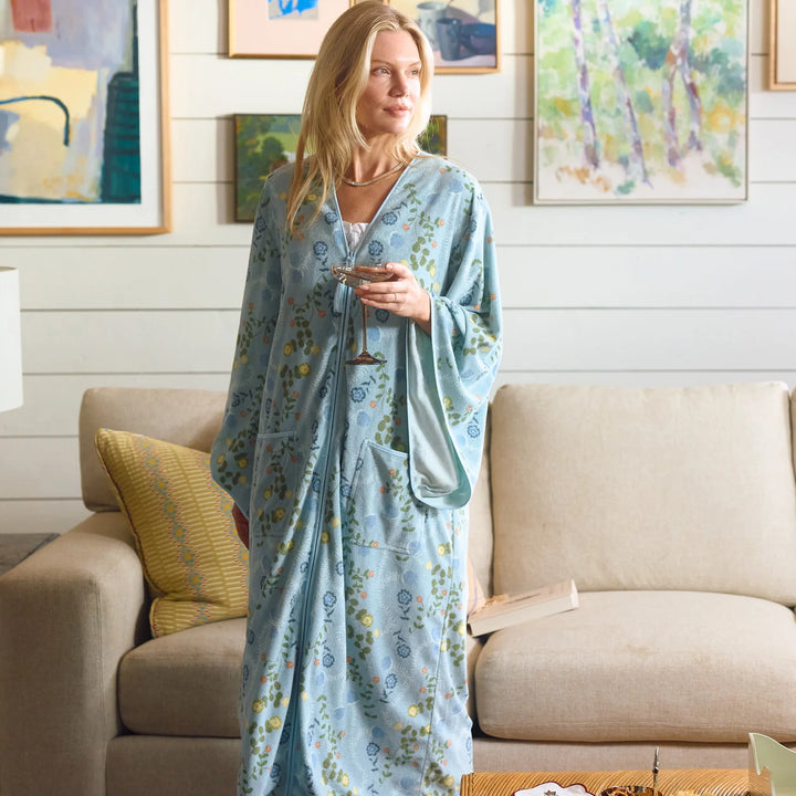 A woman wearing a light blue floral robe stands in a living room in front of a beige sofa, holding a phone.