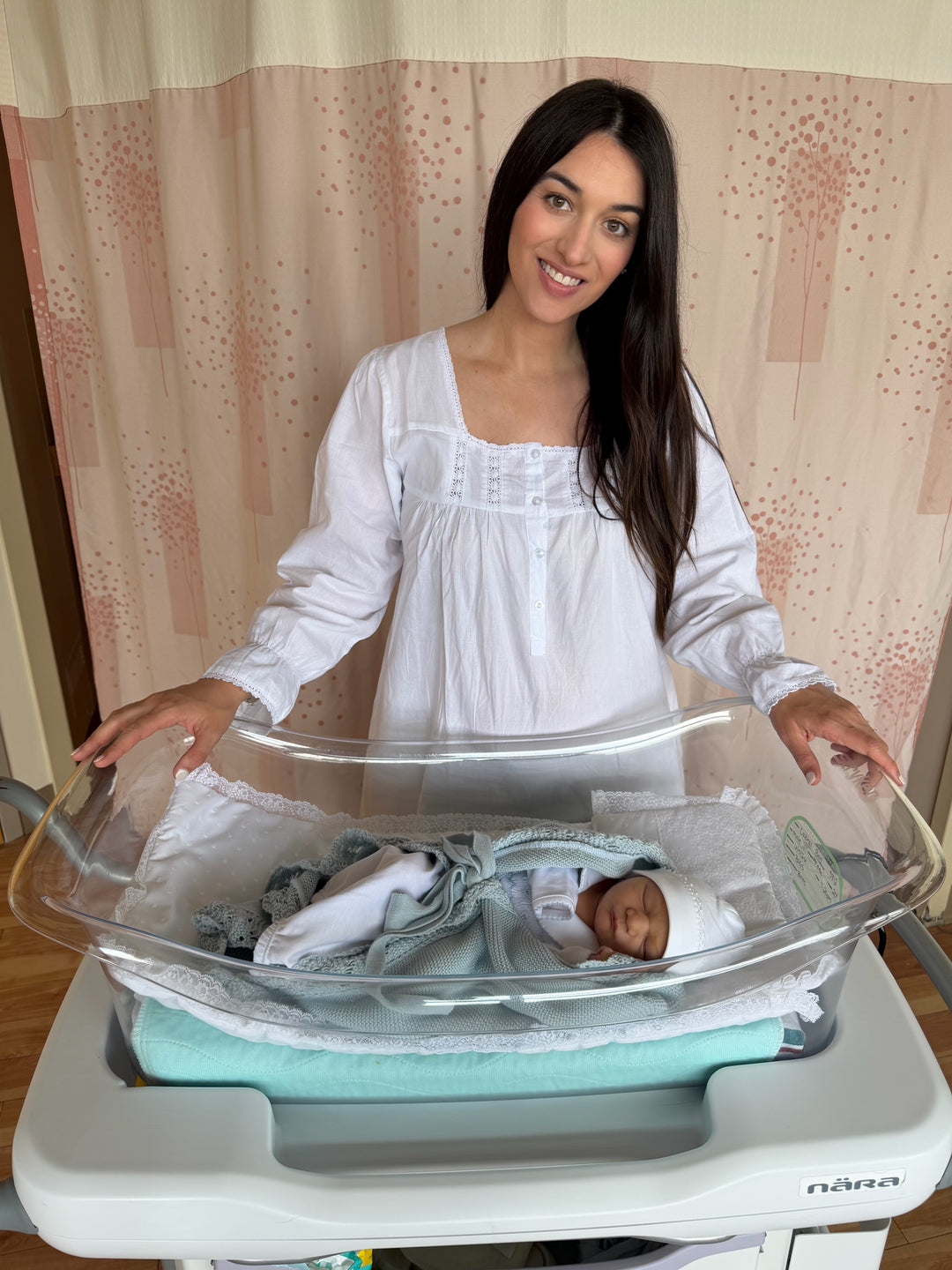 A woman in a white long-sleeve nightgown stands behind a clear newborn bassinet with a baby sleeping inside, set against a pink patterned curtain background.