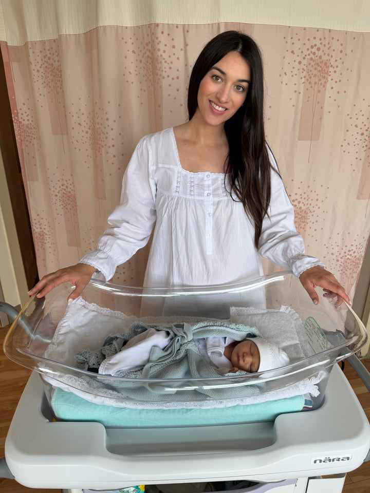 A woman in a white long-sleeve nightgown stands behind a clear newborn bassinet with a baby sleeping inside, set against a pink patterned curtain background.