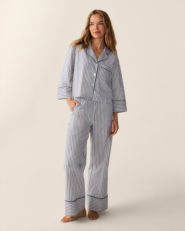 A woman modeling a matching two-piece pajama set with vertical blue and white stripes, featuring a long-sleeve button-front top and matching pants, against a plain beige studio background.