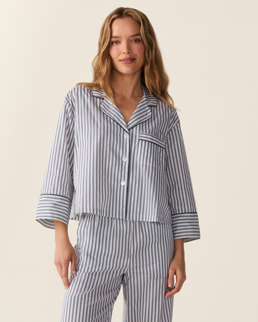 A woman modeling a matching two-piece pajama set with vertical navy and white stripes, featuring a long-sleeve button-front top and coordinating pants, against a solid light beige background.