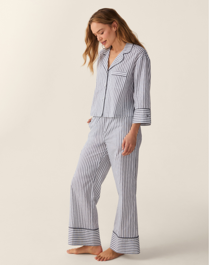 A woman modeling a matching two-piece pajama set with vertical navy and white stripes, featuring a long-sleeve button-front top and flared pants, against a plain beige studio background.