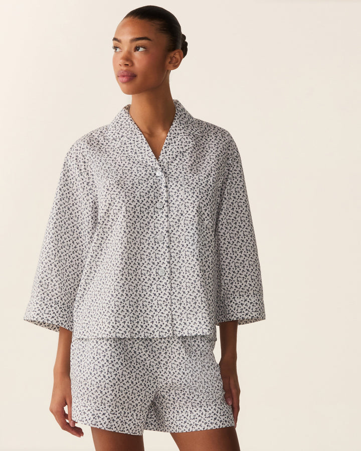 A woman modeling a matching two-piece pajama set with a white base and small black floral print, featuring a short-sleeve button-front top and shorts, against a plain beige studio background.