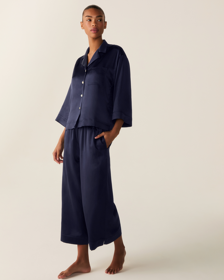 A model stands barefoot against a plain light beige studio background wearing a matching navy blue pajama set consisting of a short-sleeve button-front shirt and wide-leg cropped pants.
