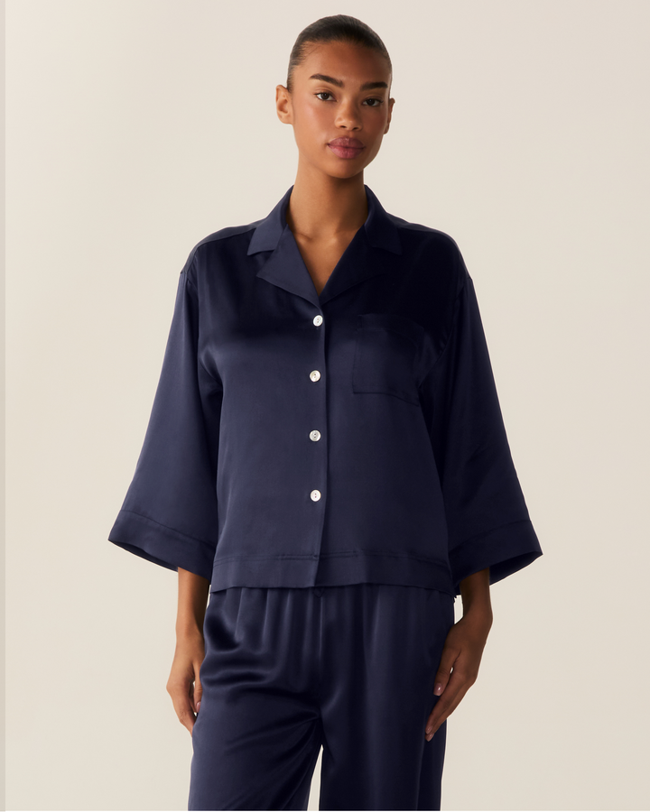 A model wearing a dark navy blue satin pajama set, featuring a short-sleeve button-up shirt and matching pants, against a solid light beige background.