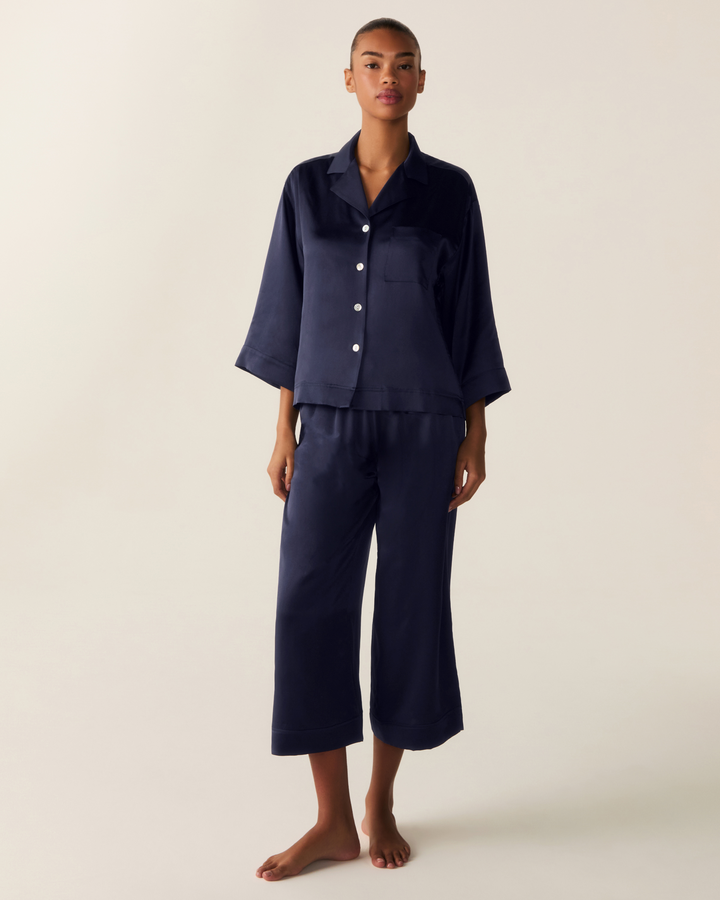A model stands barefoot wearing a matching navy blue pajama set consisting of a short-sleeve button-front top and cropped pants, photographed against a plain light beige studio background.