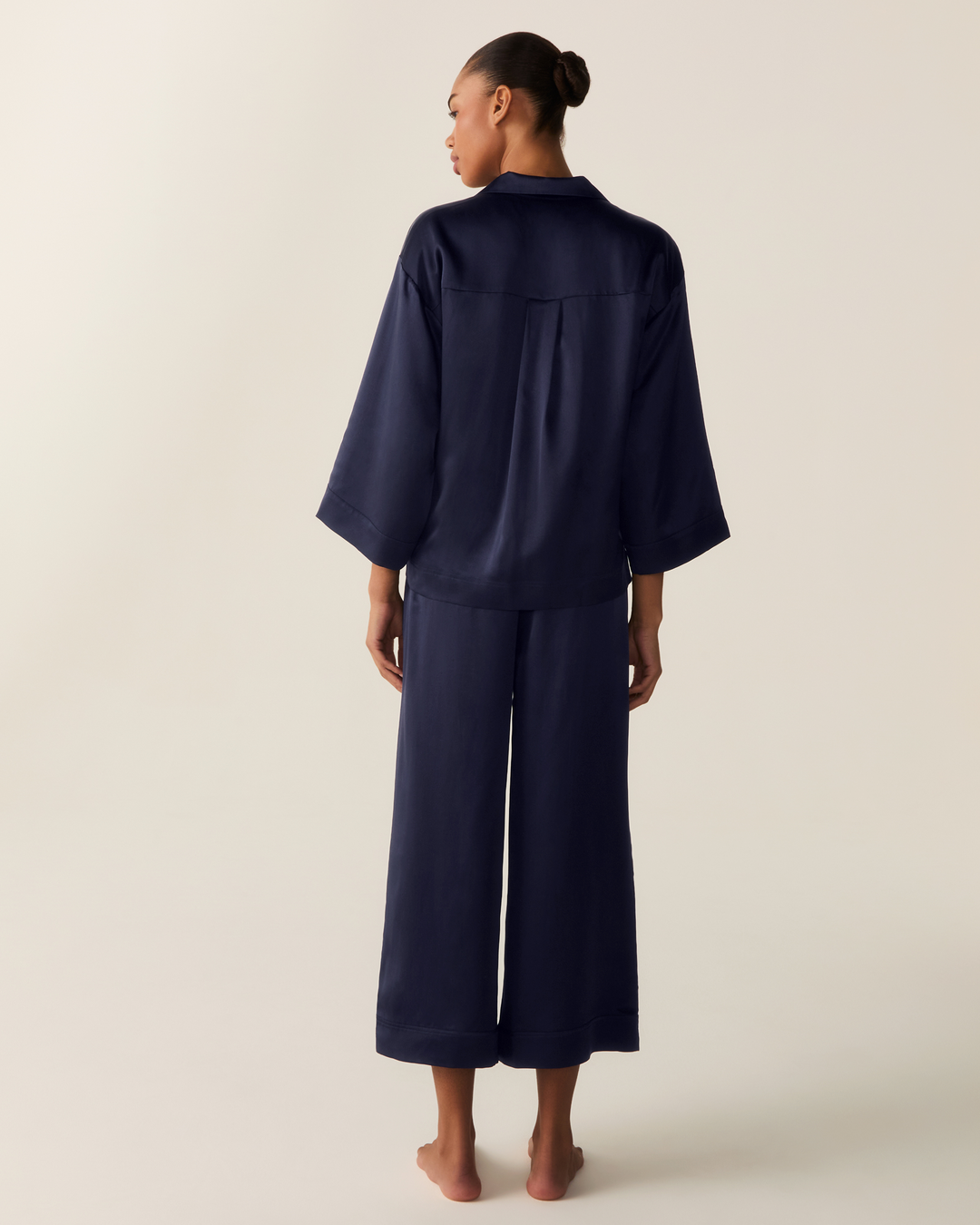 A model is shown from the back wearing a matching navy blue pajama set consisting of a short-sleeve top and wide-leg pants, against a plain light beige studio background.