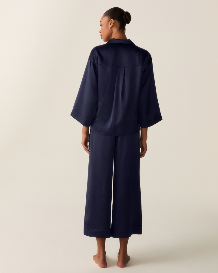 A model is shown from the back wearing a matching navy blue pajama set consisting of a short-sleeve top and wide-leg pants, against a plain light beige studio background.