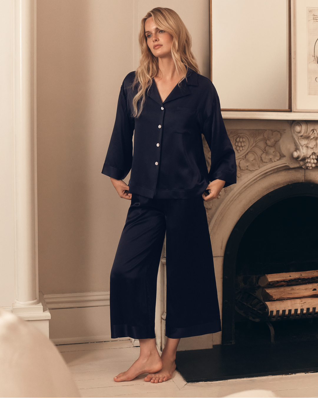 A woman stands barefoot in front of a fireplace wearing a dark navy satin pajama set consisting of a long-sleeve button-front top and matching wide-leg pants.