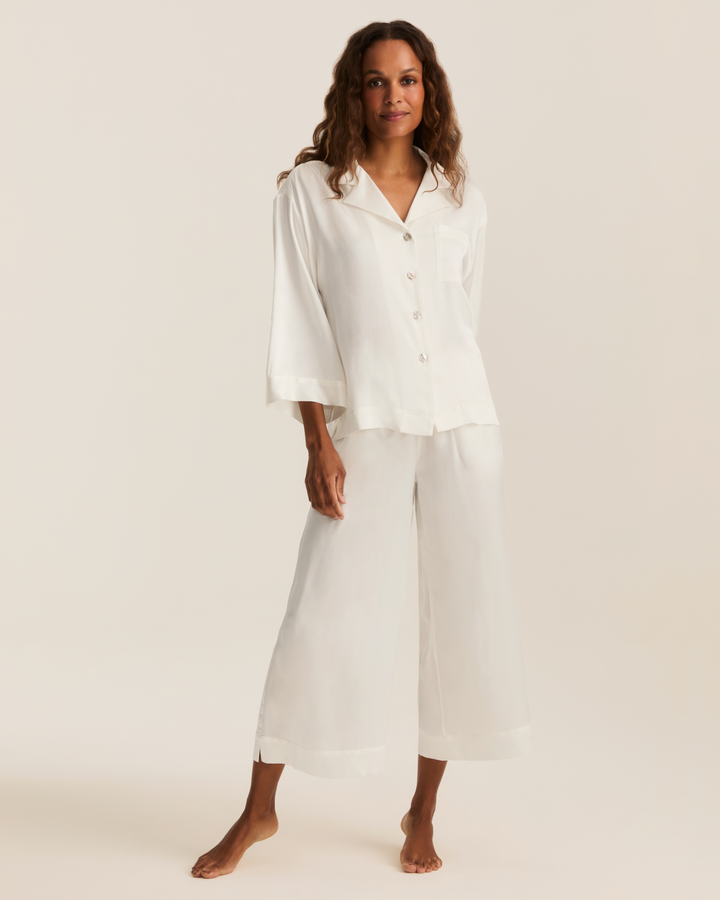 A woman modeling a white two-piece lounge set consisting of a short-sleeve button-front top and matching wide-leg cropped pants, photographed against a plain warm off-white studio background.