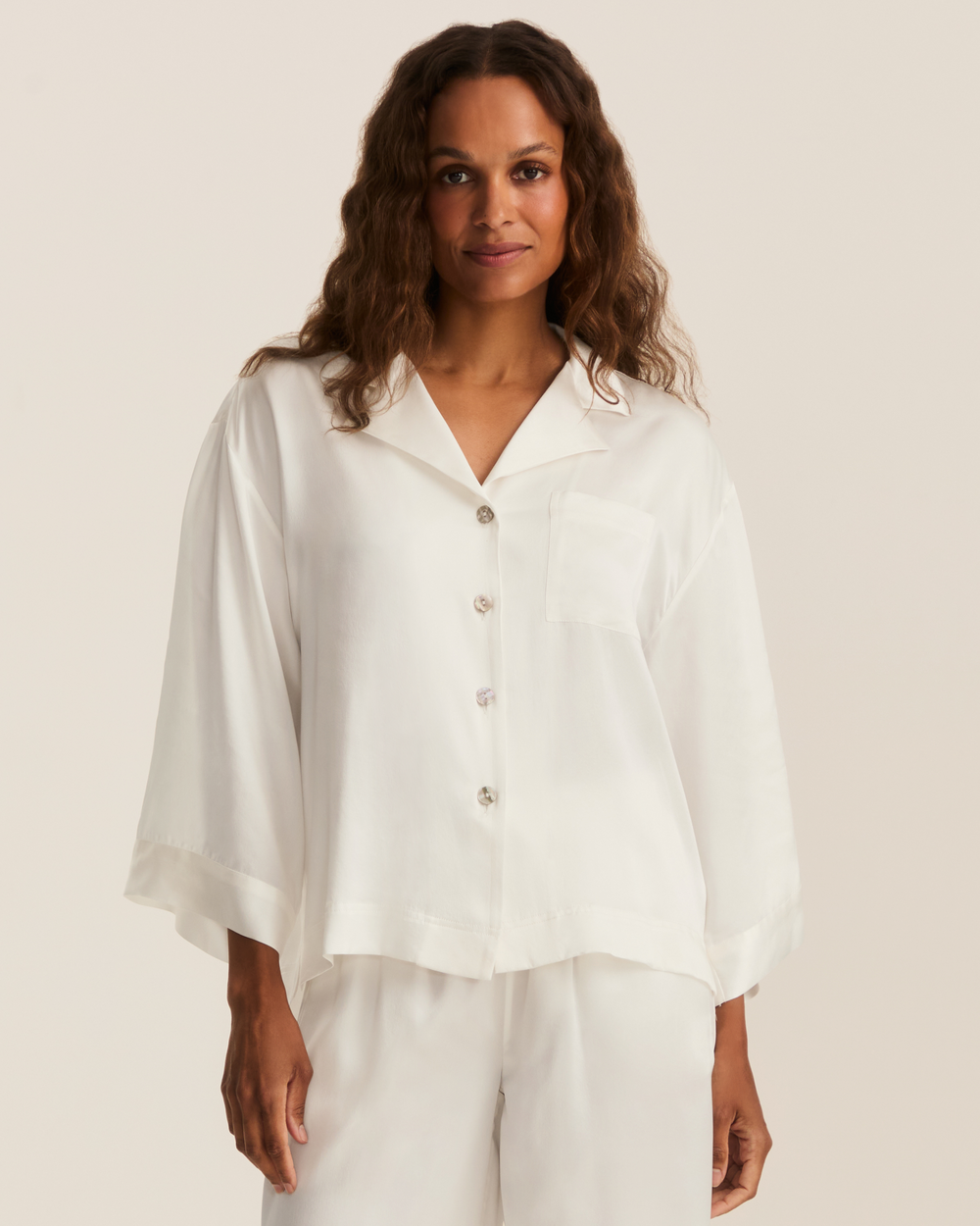 A woman wearing a white button-front short-sleeve pajama-style top with a chest pocket and matching white pants, photographed against a plain light beige studio background.