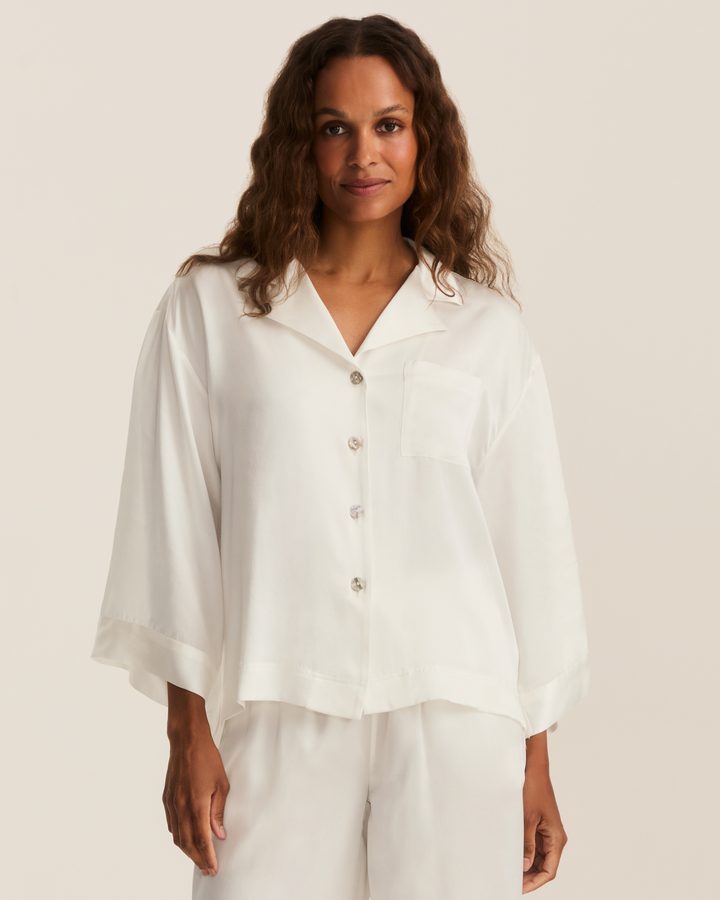 A woman wearing a white button-front short-sleeve pajama-style top with a chest pocket and matching white pants, photographed against a plain light beige studio background.