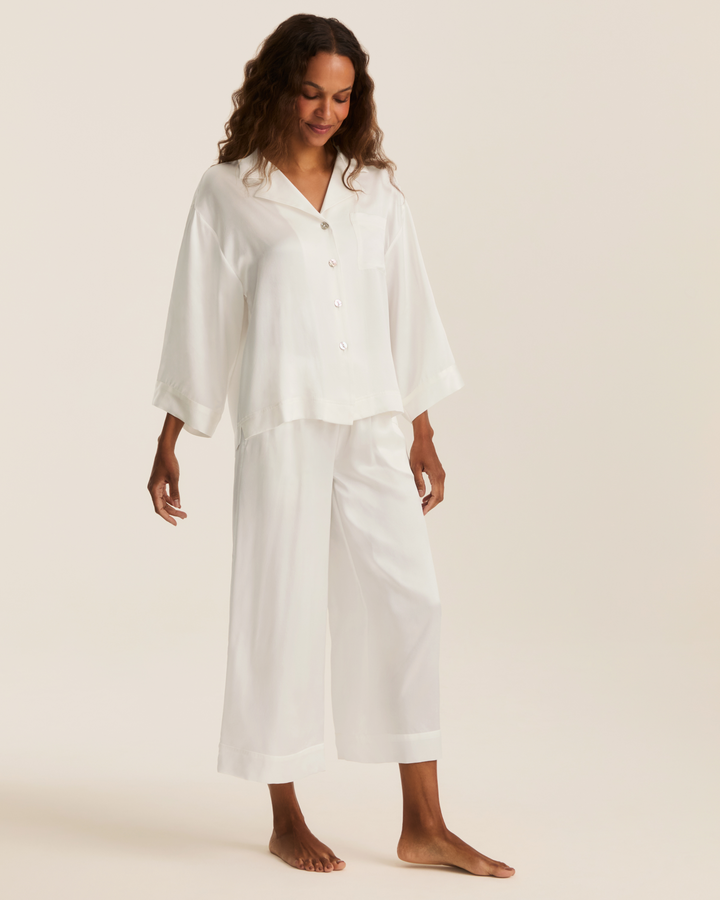 A woman modeling a white two-piece pajama set consisting of a short-sleeve button-front top and matching wide-leg pants, photographed against a plain warm off-white studio background.