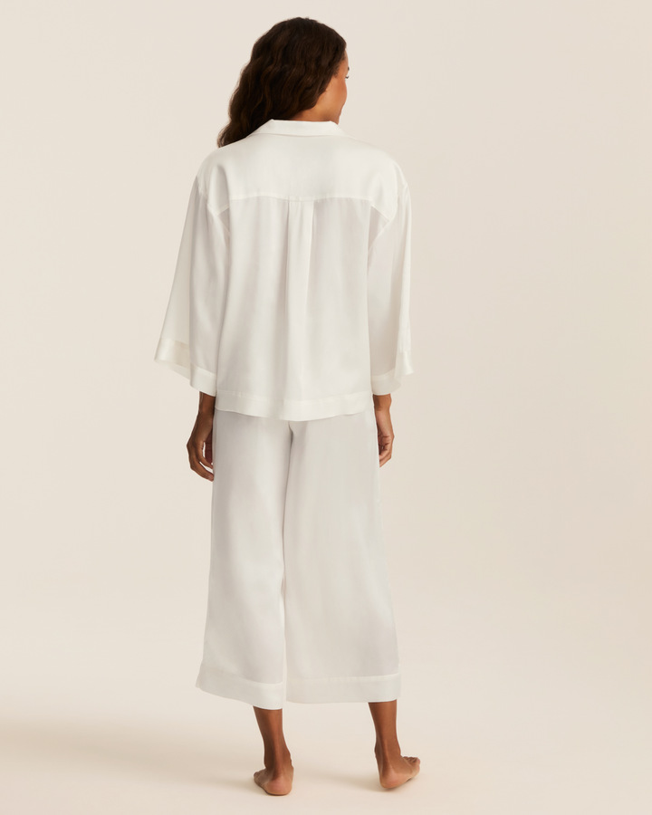 A woman stands barefoot facing away from the camera, wearing a white two-piece lounge set consisting of a short-sleeve button-up shirt and matching wide-leg cropped pants, against a solid light beige studio background.