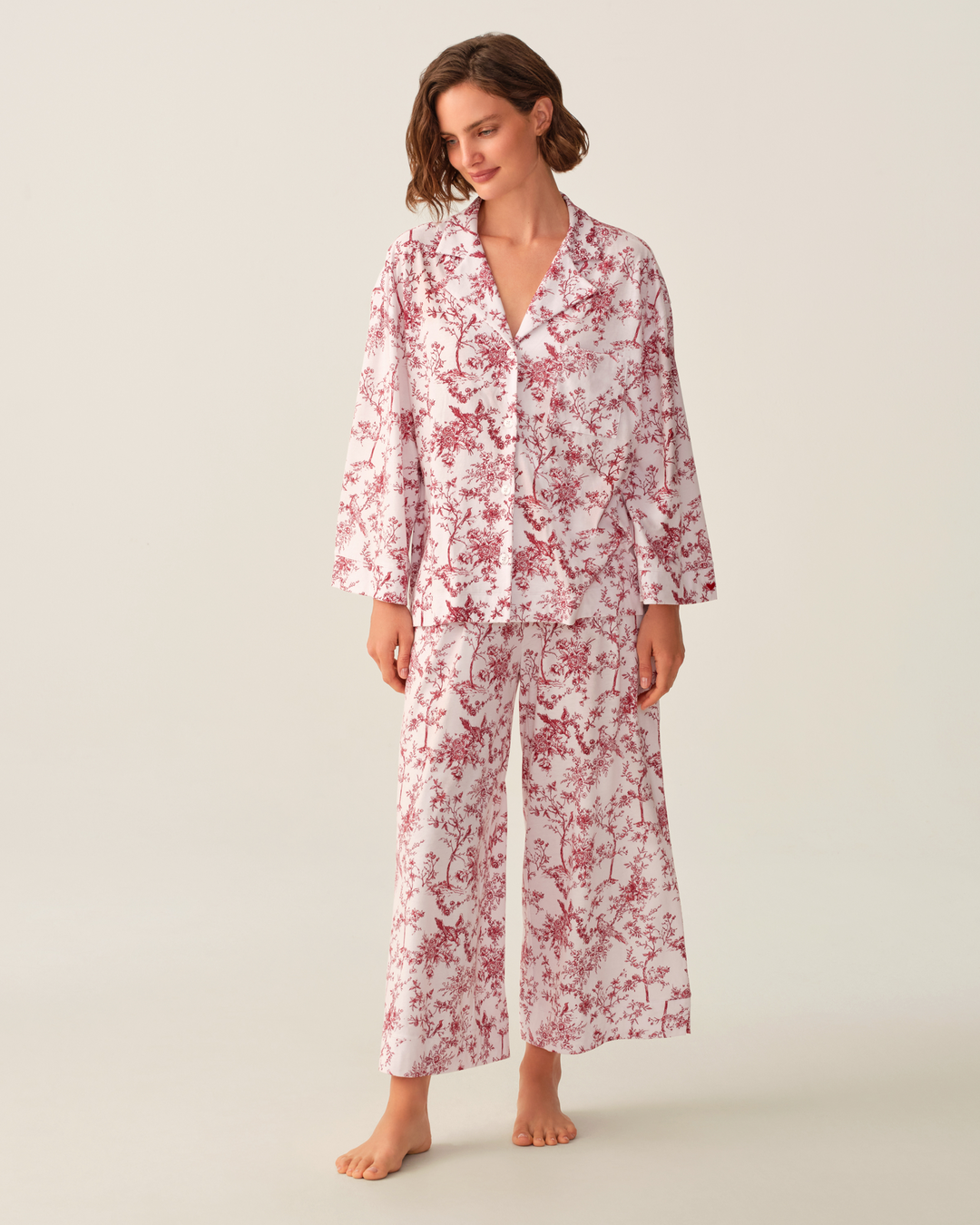 A woman modeling a matching pajama set with a pink and white floral print, consisting of a long-sleeve button-up top and wide-leg pants, against a solid light beige background.