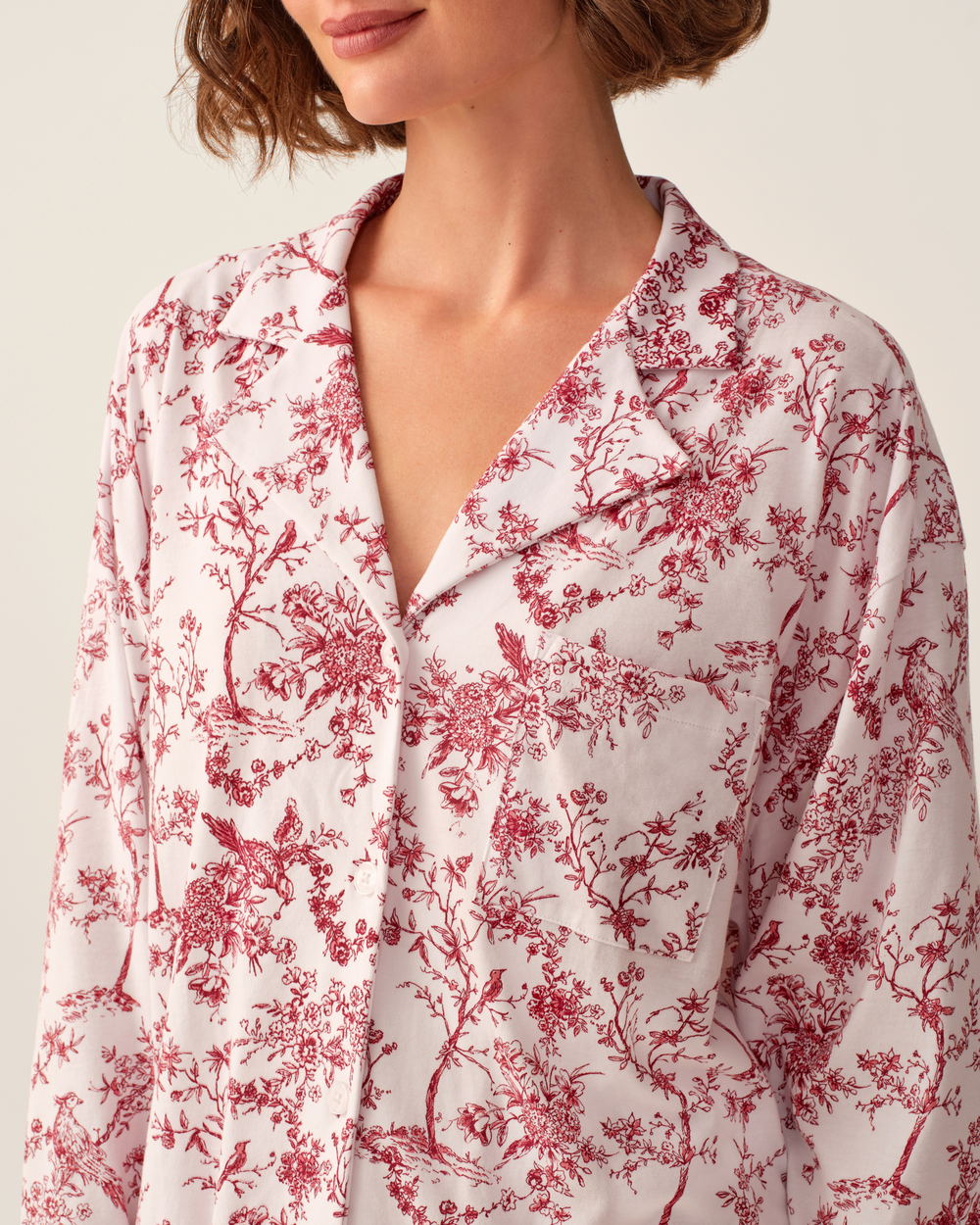 A close-up of a model wearing a long-sleeve collared button-front shirt with a red toile floral and bird pattern on a white background.