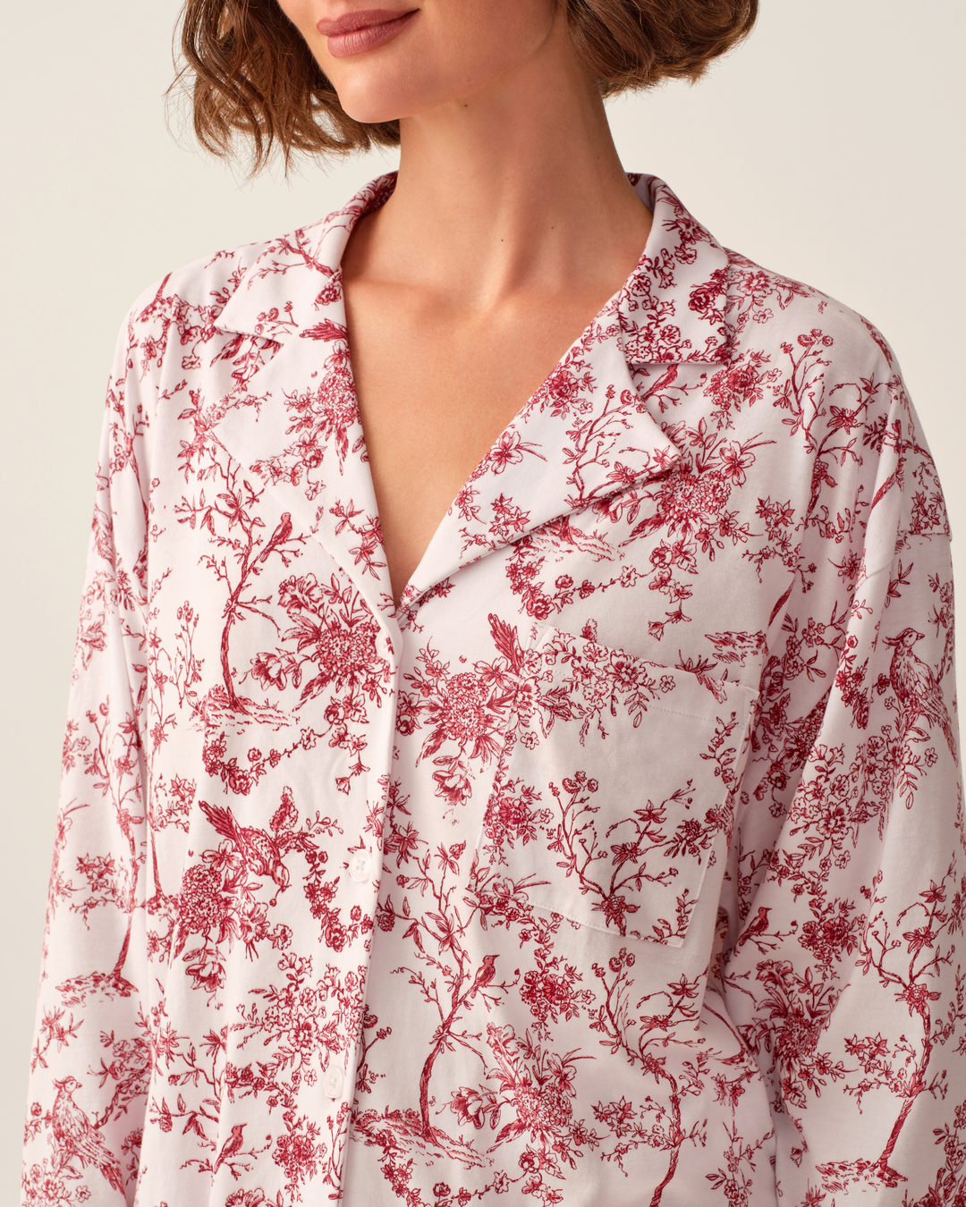 A close-up of a model wearing a long-sleeve collared button-front shirt with a red toile floral and bird pattern on a white background.