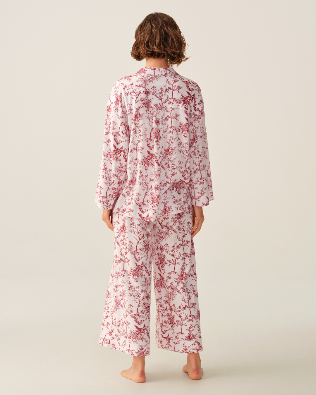 A model viewed from the back wearing a matching two-piece pajama set with a red floral print on a white background, set against a solid light beige studio backdrop.