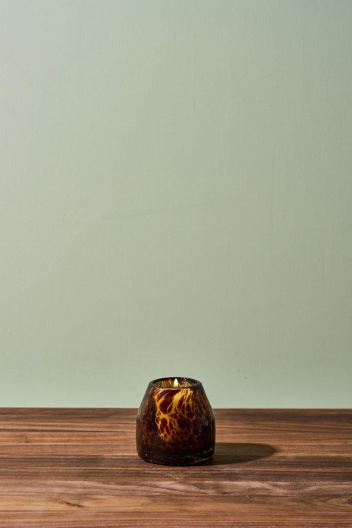 A small amber glass candle holder with a marbled, swirled pattern sits on a dark wood surface against a solid sage green background.