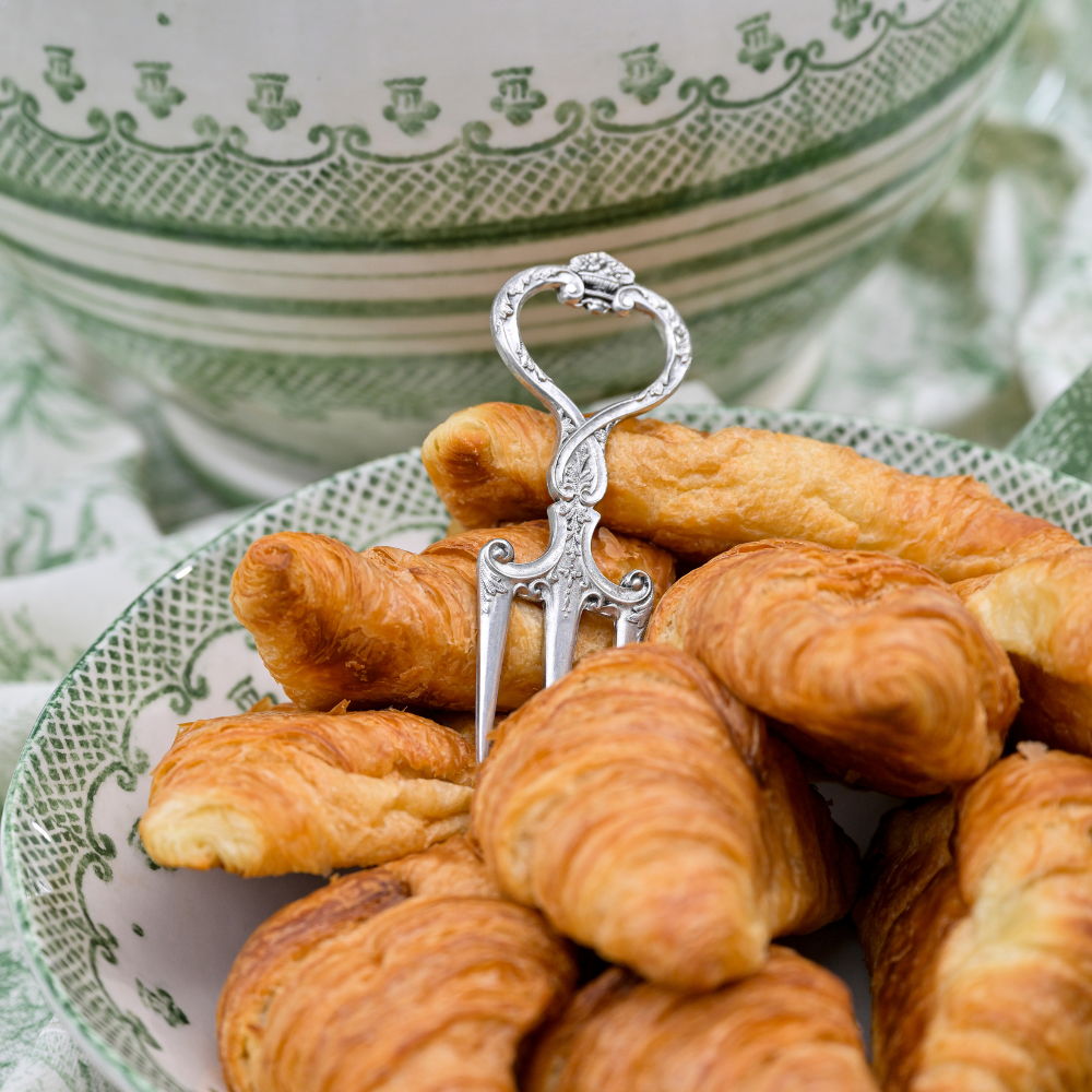 Silver pastry fork on a plate of croissants with a decorative green and white bowl in the background.