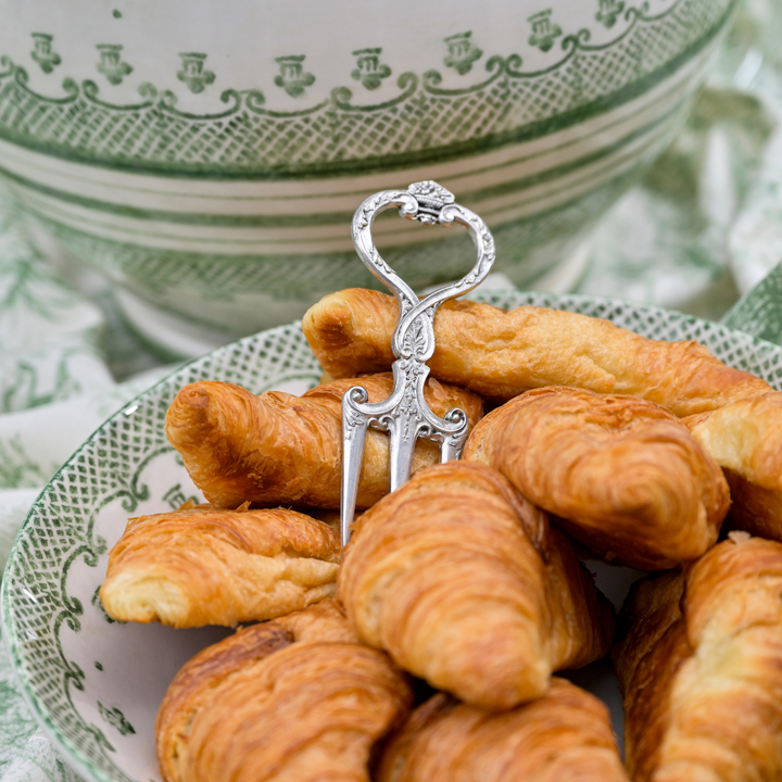 Silver pastry fork on a plate of croissants with a decorative green and white bowl in the background.
