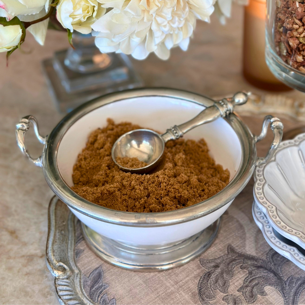 White bowl with brown powder and a silver scoop on a decorative tablecloth.